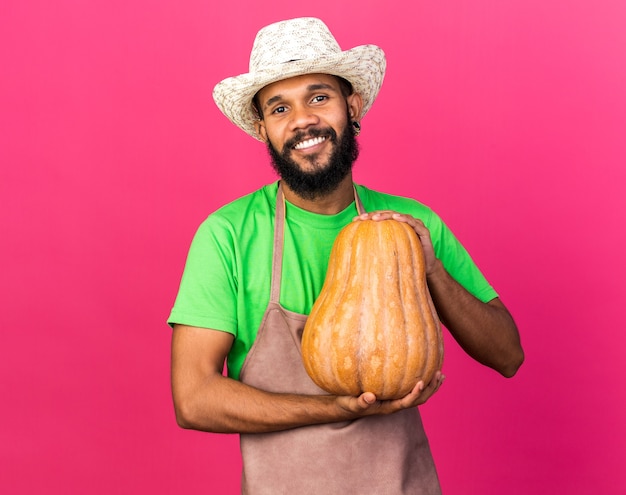 Lächelnder junger afroamerikanischer gärtner mit gartenhut, der kürbis isoliert auf rosa wand hält