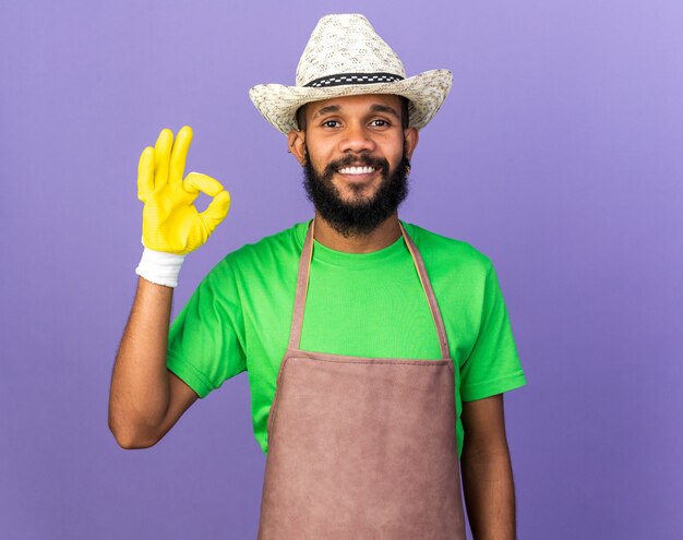 Lächelnder junger afroamerikanischer Gärtner mit Gartenhut mit Handschuhen, der eine gute Geste einzeln auf blauer Wand zeigt
