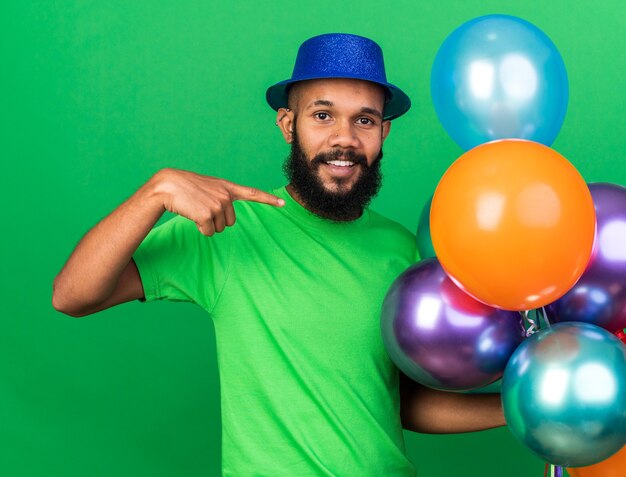 Lächelnder junger afroamerikanischer Kerl mit Partyhut, der auf Luftballons zeigt und zeigt