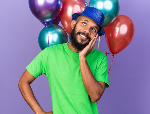 Lächelnder junger afroamerikanischer Kerl mit Partyhut, der vor Ballons steht und Hand auf das Kinn legt