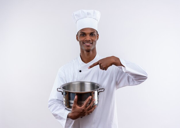 Lächelnder junger afroamerikanischer Koch in der Kochuniform hält Topf und Punkte am Topf auf lokalisiertem weißem Hintergrund mit Kopienraum