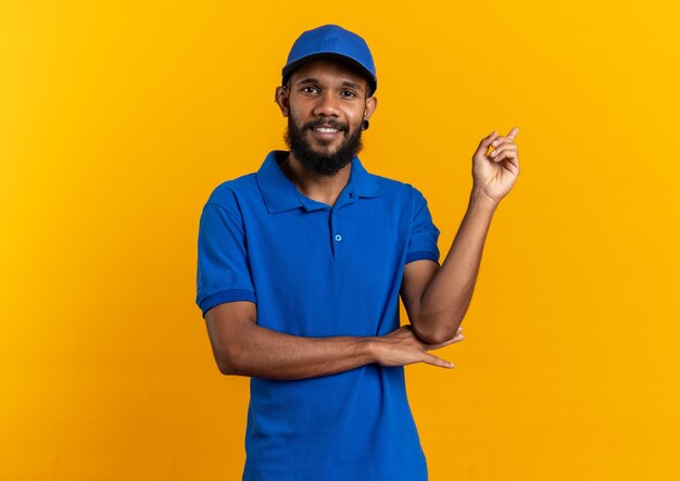 Lächelnder junger afroamerikanischer Lieferbote, der auf die Seite zeigt, die auf orangefarbenem Hintergrund mit Kopienraum isoliert ist?