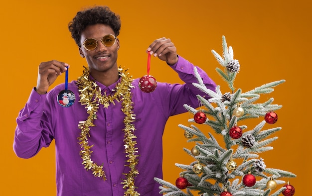 Lächelnder junger afroamerikanischer Mann, der Brille mit Lametta-Girlande um den Hals trägt, der nahe verziertem Weihnachtsbaum auf orange Hintergrund steht