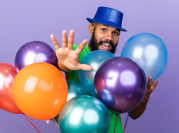Lächelnder junger afroamerikanischer Mann mit Partyhut, der hinter Ballons steht und die Hand vorne isoliert auf blauer Wand hält