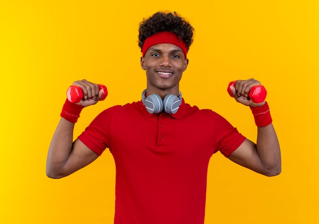 Lächelnder junger afroamerikanischer sportlicher Mann, der Stirnband und Armband in Kopfhörern am Hals trägt, der Hanteln an der gelben Wand anhebt
