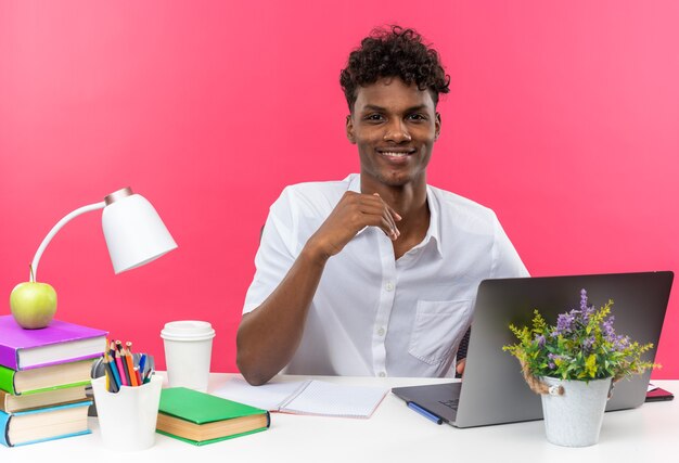 Lächelnder junger afroamerikanischer Student, der am Schreibtisch mit Schulwerkzeugen sitzt, die isoliert auf rosa Wand aussehen?