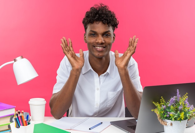 Lächelnder junger afroamerikanischer Student, der am Schreibtisch mit Schulwerkzeugen sitzt und die Hände offen hält