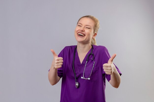 Lächelnder junger Arzt, der lila medizinisches Kleid trägt und ihre Daumen auf isolierte weiße Wand stethoskopiert