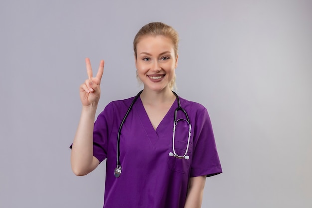 Lächelnder junger Arzt, der lila medizinisches Kleid und Stethoskop trägt, zeigt Friedensgeste auf isolierter weißer Wand