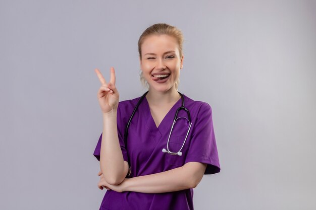 Lächelnder junger Arzt, der lila medizinisches Kleid und Stethoskop trägt, zeigt Friedensgeste auf isolierter weißer Wand