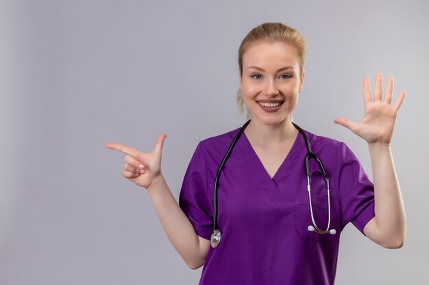 Lächelnder junger Arzt, der lila medizinisches Kleid und Stethoskop trägt, zeigt unterschiedliche Geste auf isolierter weißer Wand