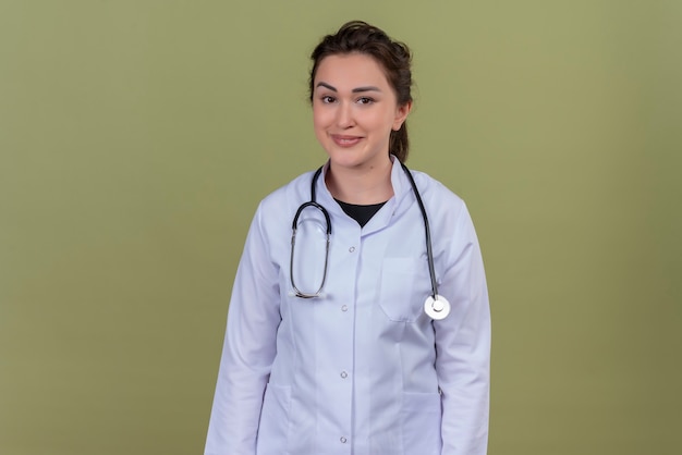 Lächelnder junger Arzt, der medizinisches Kleid trägt, das Stethoskop auf grüner Wand trägt