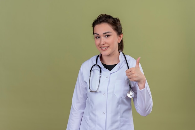Lächelnder junger Arzt, der medizinisches Kleid trägt, das Stethoskop ihren Daumen oben auf grüner Wand trägt