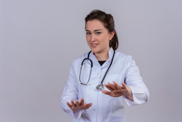 Lächelnder junger Arzt, der medizinisches Kleid trägt, das Stethoskop trägt, zeigt Größe auf weißer Wand