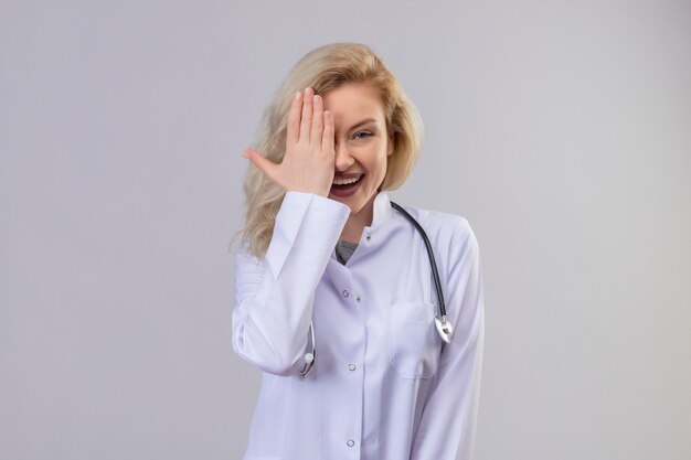 Lächelnder junger Arzt, der Stethoskop im medizinischen Kleid trägt, bedeckte Auge auf weißer Wand