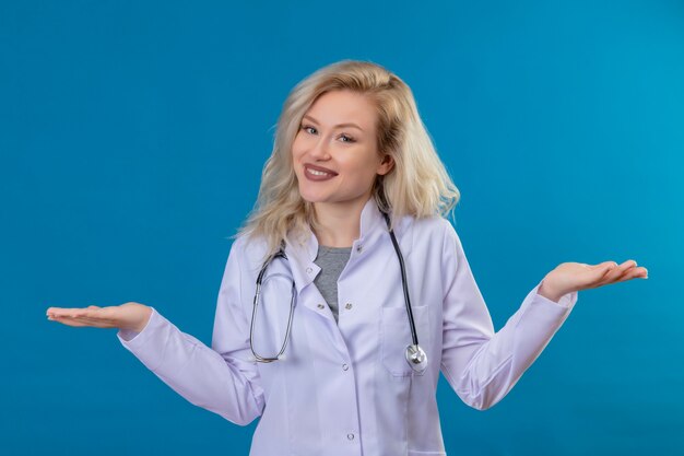 Lächelnder junger Arzt, der Stethoskop im medizinischen Kleid trägt, breitet Hände auf blauer Wand aus