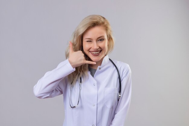 Lächelnder junger Arzt, der Stethoskop im medizinischen Kleid trägt, das Anrufgeste auf weißer Wand zeigt
