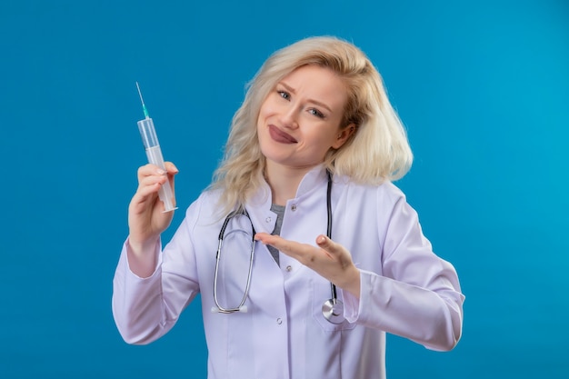 Lächelnder junger Arzt, der Stethoskop im medizinischen Kleid trägt Spritze auf blauer Wand hält