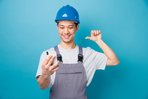 Lächelnder junger Bauarbeiter, der Schutzhelm und Uniform hält, hält und betrachtet Handy, das auf sich selbst zeigt