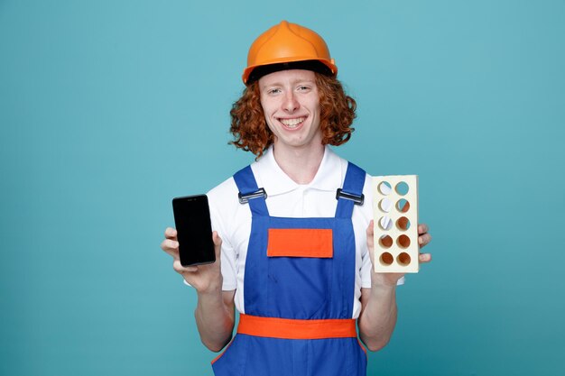 Lächelnder junger Baumeister in Uniform, der Telefon mit Ziegeln isoliert auf blauem Hintergrund hält