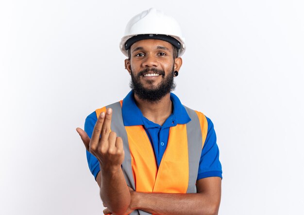 Lächelnder junger Baumeister in Uniform mit Schutzhelm, der jemanden mit der Hand anruft, isoliert auf weißer Wand mit Kopierraum