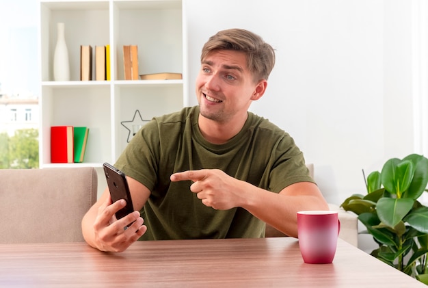 Lächelnder junger blonder gutaussehender mann sitzt am tisch mit der tasse, die hält und auf telefon zeigt, das seite innerhalb des wohnzimmers schaut