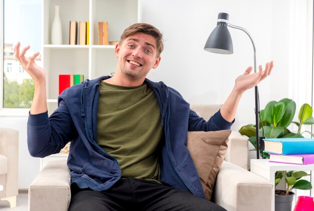Lächelnder junger blonder hübscher Mann sitzt auf Sessel und betrachtet Kamera mit erhobenen Händen innerhalb des Wohnzimmers