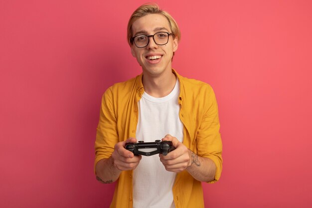 Lächelnder junger blonder Kerl, der gelbes T-Shirt und Brillen-Gamecontroller-Joystick trägt, der auf Rosa lokalisiert wird