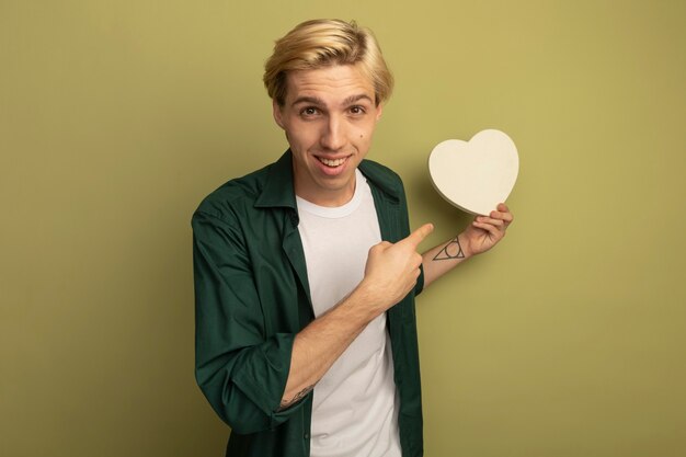 Lächelnder junger blonder Kerl, der grünes T-Shirt hält und auf Herzformbox zeigt