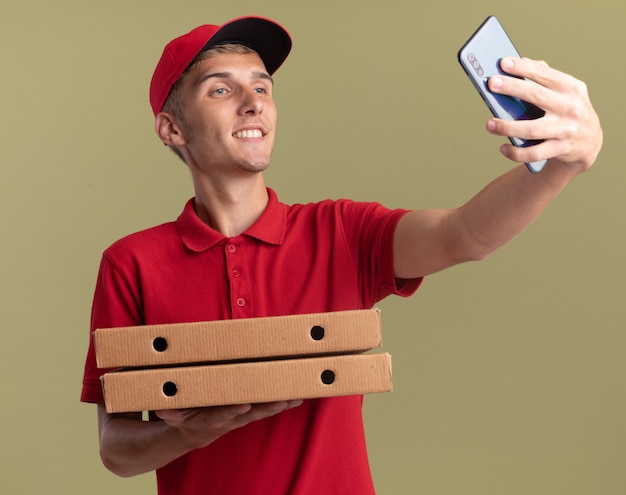 Lächelnder junger blonder Lieferjunge hält Pizzakartons und sieht sich das Telefon an, das ein Selfie macht