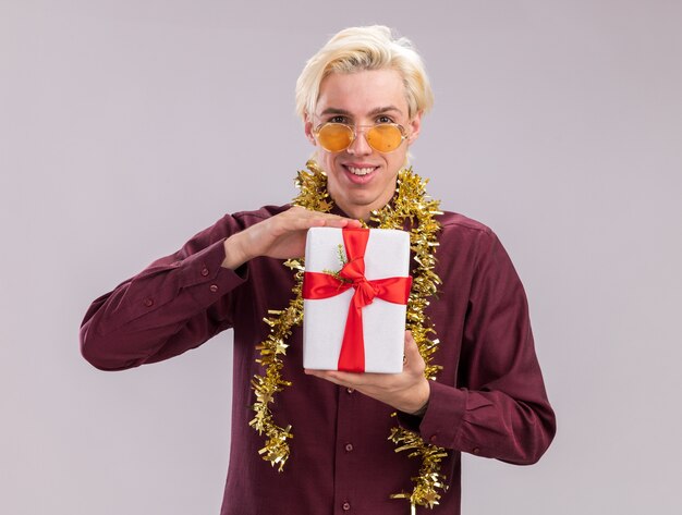 Lächelnder junger blonder Mann, der Brille mit Lametta-Girlande um den Hals hält, der Geschenkpaket hält, Kamera betrachtet auf weißem Hintergrund