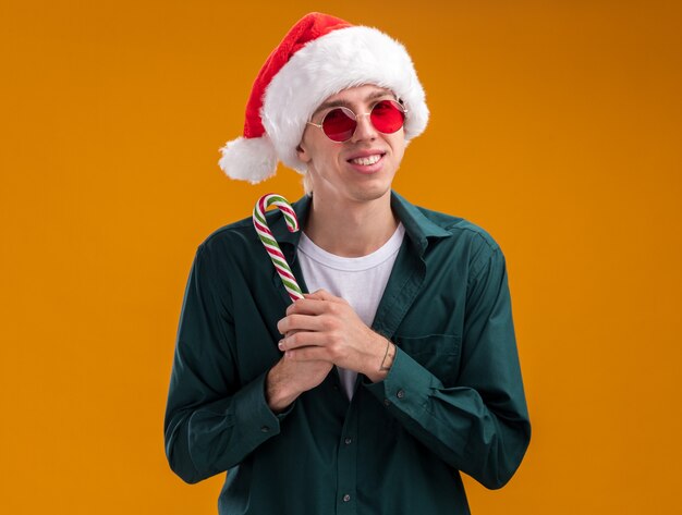 Lächelnder junger blonder Mann mit Weihnachtsmütze und Brille, der weihnachtlichen Zuckerrohr hält und die Kamera einzeln auf orangefarbenem Hintergrund betrachtet