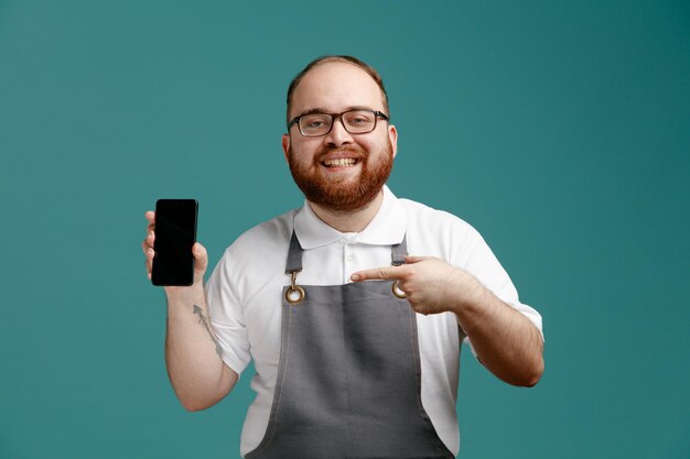 Lächelnder junger Friseur in Uniform und Brille, der in die Kamera blickt und ein Handy zeigt, das darauf isoliert auf blauem Hintergrund zeigt