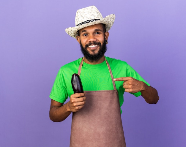 Lächelnder junger Gärtner afroamerikanischer Mann mit Gartenhut hält und zeigt auf Aubergine