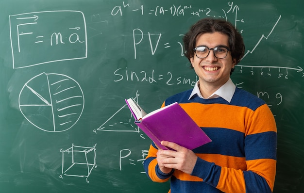 Lächelnder junger Geometrielehrer mit Brille, der im Klassenzimmer vor einer Tafel steht und ein offenes Buch hält, das nach vorne schaut