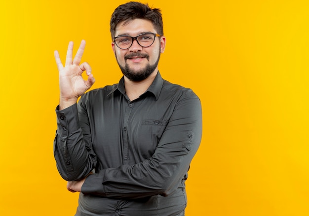 Lächelnder junger Geschäftsmann, der Brillen trägt, die okey Geste lokalisiert auf Gelb zeigen