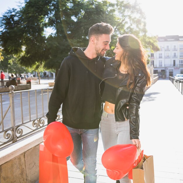 Lächelnder junger glücklicher Kerl Dame mit Paketen und Ballonen auf Straße umfassend