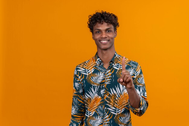 Lächelnder junger gutaussehender dunkelhäutiger Mann mit lockigem Haar im bedruckten Hemd der Blätter, das mit Zeigefinger auf einem orangefarbenen Hintergrund zeigt