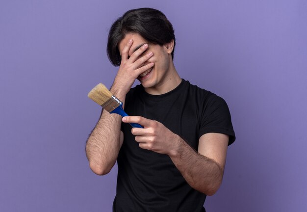 Lächelnder junger gutaussehender Kerl, der ein schwarzes T-Shirt trägt, das Pinsel bedecktes Gesicht mit der Hand hält, die auf purpurroter Wand isoliert ist?