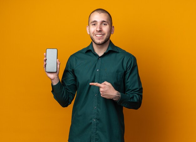 Lächelnder junger gutaussehender Kerl, der grünes Hemd hält und Punkte auf Telefon trägt