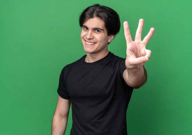 Lächelnder junger gutaussehender Kerl mit schwarzem T-Shirt, das drei isoliert auf grüner Wand zeigt
