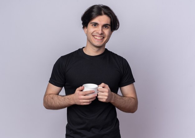 Lächelnder junger gutaussehender Kerl mit schwarzem T-Shirt mit Tasse Kaffee isoliert auf weißer Wand