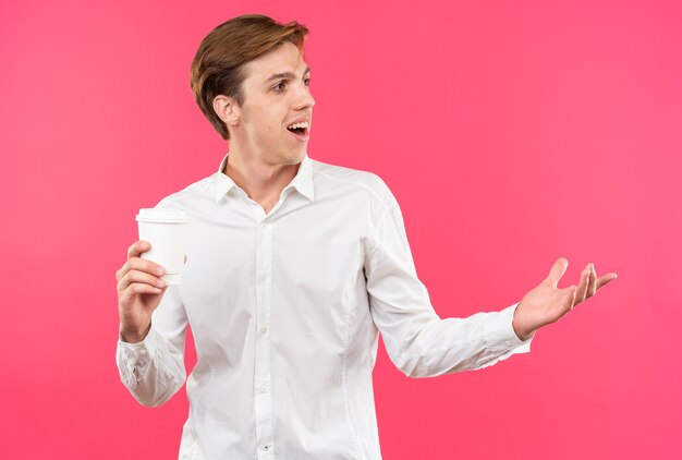 Lächelnder junger gutaussehender Kerl mit weißem Hemd, der eine Tasse Kaffee mit der Hand an der Seite hält, isoliert auf rosa Wand