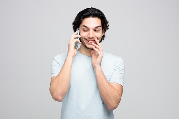 Lächelnder junger gutaussehender Mann, der am Telefon spricht und die Hand in der Nähe des Mundes hält, während er das Flüstern der Seite isoliert auf weißem Hintergrund betrachtet