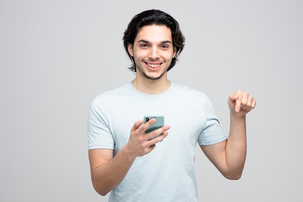 Lächelnder junger gutaussehender Mann, der das Handy hält und in die Kamera schaut, die nach unten zeigt, isoliert auf weißem Hintergrund