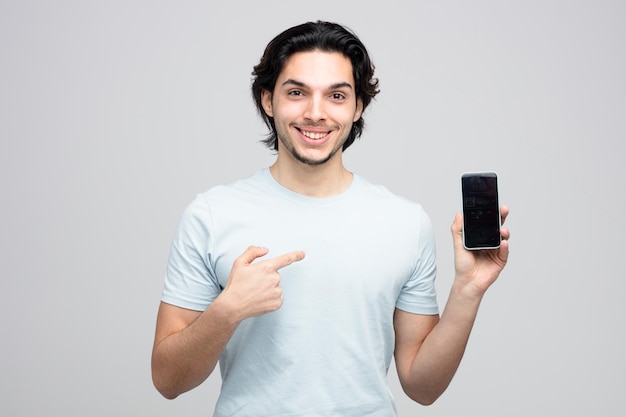 Lächelnder junger gutaussehender Mann, der das Handy zeigt, das darauf zeigt und die Kamera isoliert auf weißem Hintergrund betrachtet