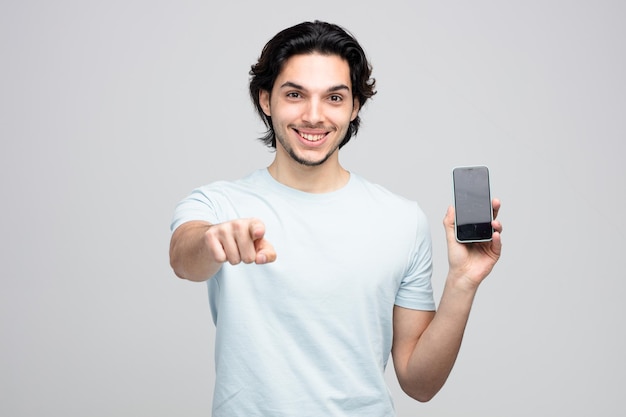 lächelnder junger gutaussehender Mann, der das Handy zeigt und auf die Kamera zeigt, die auf weißem Hintergrund isoliert ist