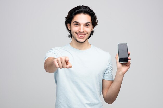 lächelnder junger gutaussehender Mann, der das Handy zeigt und auf die Kamera zeigt, die auf weißem Hintergrund isoliert ist