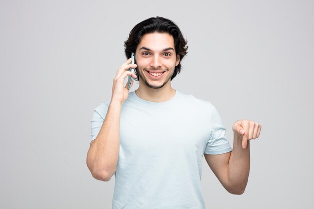 Lächelnder junger gutaussehender Mann, der die Kamera ansieht, die nach unten zeigt, während er am Telefon spricht, isoliert auf weißem Hintergrund