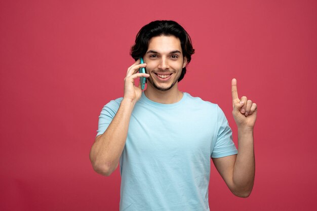 lächelnder junger gutaussehender Mann, der in die Kamera blickt und am Telefon nach oben zeigt, isoliert auf rotem Hintergrund